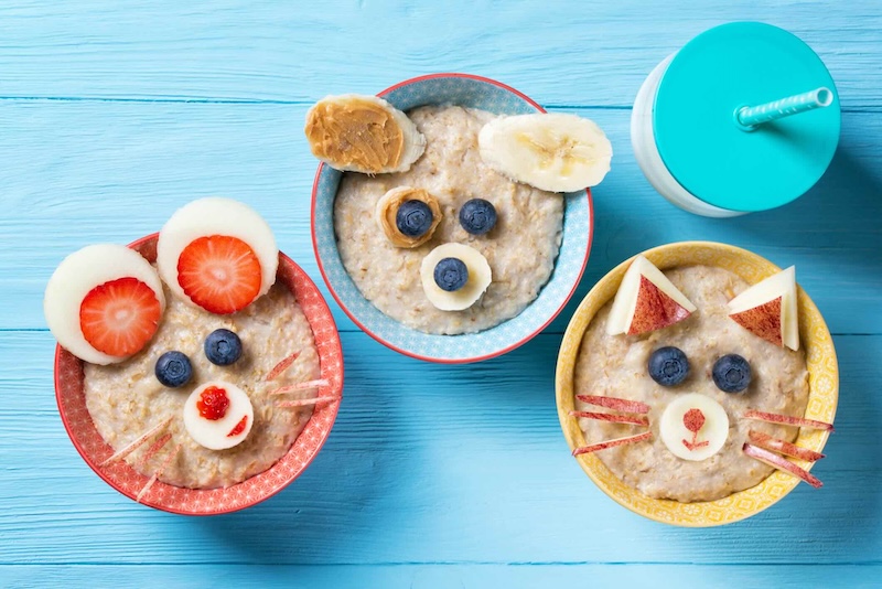 Funny bowls with oat porridge with cat, dog and mouse faces made of fruits and berries, food for kids idea, top view